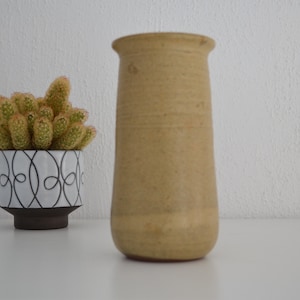 May include: A light brown ceramic vase with a rounded bottom and a slightly flared top. The vase is sitting on a white surface.
