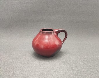 May include: A small, red ceramic vase with a rounded body and a single handle. The vase has a dark glaze on the bottom.