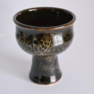 Peut inclure: Un vase en céramique brun foncé avec un glaçage moucheté. Le vase a un large bol rond et une tige haute et mince.