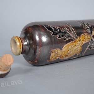 May include: A brown ceramic bottle with a cork stopper. The bottle is decorated with a stylised bird and floral design in shades of brown, yellow, and black.