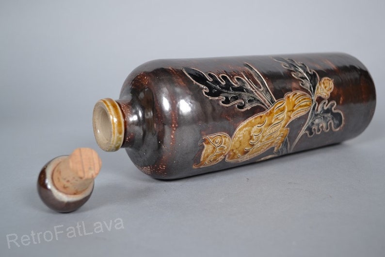 May include: A brown ceramic bottle with a cork stopper. The bottle is decorated with a stylised bird and floral design in shades of brown, yellow, and black.