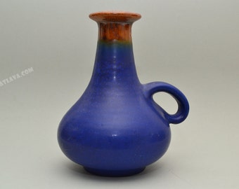 Puede incluir: Un jarrón de cerámica azul con un borde marrón y un asa. El jarrón tiene un cuerpo redondeado y un cuello estrecho.