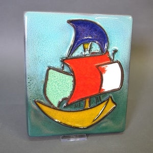 Op de afbeelding: Een keramische tegel met een kleurrijke illustratie van een zeilboot met een blauw zeil, een rood en wit lichaam en een gele romp. De tegel heeft een groene achtergrond.