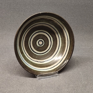 Puede incluir: Un cuenco de cerámica con círculos concéntricos en tonos marrón, blanco y verde. El cuenco tiene un exterior marrón oscuro y un acabado brillante. El diseño presenta una serie de anillos que irradian desde el centro.