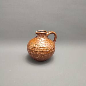 May include: A small, brown ceramic jug with a textured surface. The jug has a round body and a short, curved handle.