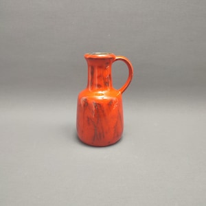 Op de afbeelding: Een kleine oranje keramische kan met een bruine glazuur en een zilveren rand. De kan heeft een handvat en een rond lichaam.