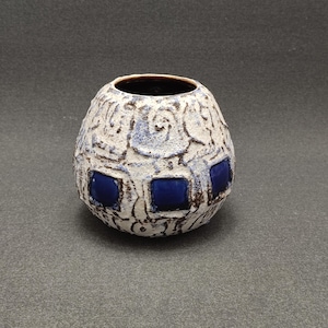 Puede incluir: Un jarrón de cerámica blanca con una superficie texturizada y detalles azules. El jarrón tiene una forma redondeada y una abertura ancha.