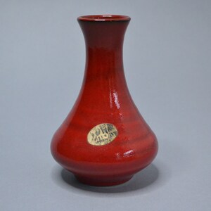 May include: A small, red ceramic vase with a round body and a narrow neck. The vase has a label on the side that reads "Studio Gallery".