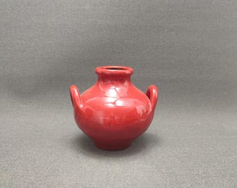 May include: A small, red ceramic vase with two handles. The vase has a rounded body and a slightly flared lip.