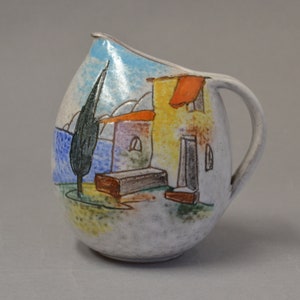 Puede incluir: Una jarra de cerámica blanca con una escena pintada a mano de una casa con un techo rojo, un árbol y un cielo azul. La jarra tiene una asa y un cuerpo redondeado.