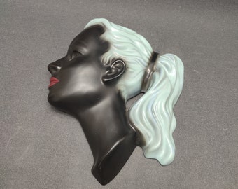 Puede incluir: Una escultura de pared de cerámica de la cabeza de una mujer con piel negra y cabello azul verdoso. El cabello de la mujer está recogido en una coleta y tiene labios rojos.