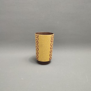 Może przedstawiać: Żółto-brązowa szklanka do picia z ozdobnym wzorem czerwonych pętli wokół środka.