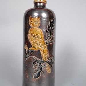 May include: A brown ceramic bottle with a brown and gold owl design. The owl is perched on a branch with leaves. The bottle has a brown ceramic stopper.