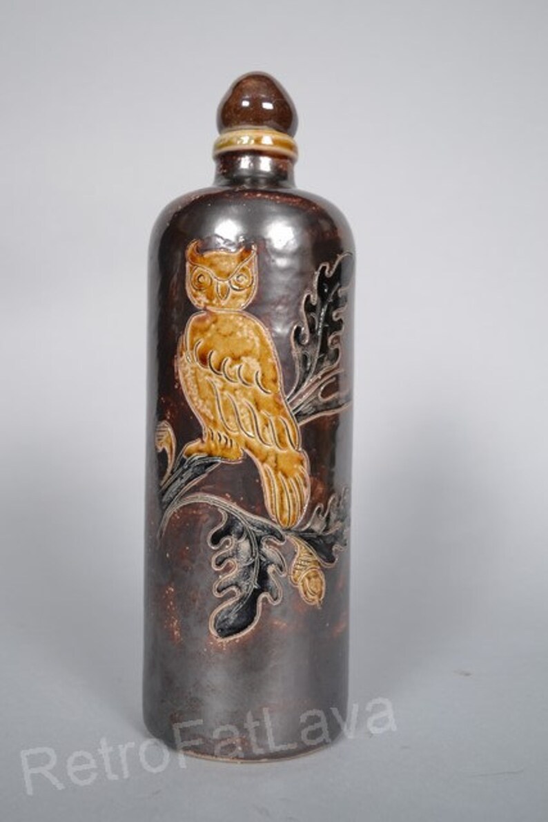 May include: A brown ceramic bottle with a brown and gold owl design. The owl is perched on a branch with leaves. The bottle has a brown ceramic stopper.