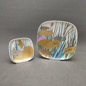 Könnte beinhalten: Zwei quadratische dekorative Teller mit weißem Hintergrund. Die Teller zeigen ein goldenes, braunes und graues Lotusblatt-Design mit rosa Lotusblüten und blauen und grünen Schilf. Die Teller werden auf transparenten Ständern präsentiert.