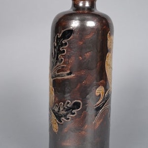 May include: A brown ceramic bottle with a decorative floral design in black and gold. The bottle has a round, brown stopper.