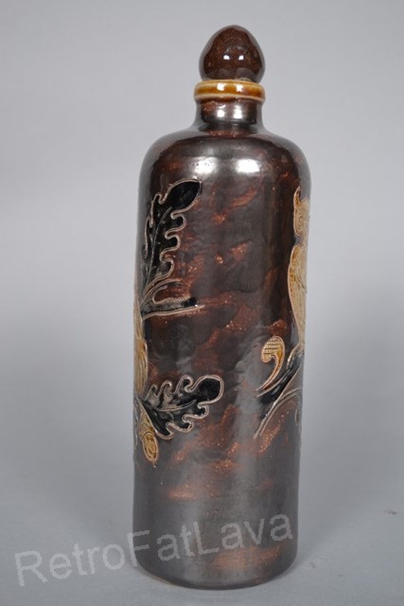 May include: A brown ceramic bottle with a decorative floral design in black and gold. The bottle has a round, brown stopper.
