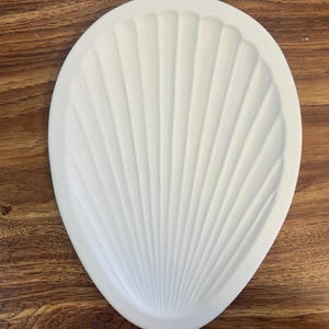 May include: A matte white, oval-shaped tray featuring a textured, radial ribbed pattern that mimics the appearance of a seashell. The object has a smooth, clean finish with a defined border and a concave center, presenting a simple, functional form.