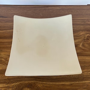 May include: A square, off-white ceramic plate with slightly curved sides. The surface features a smooth, matte finish with subtle, light brown circular stains and discoloration marks visible on the left side of the center area.