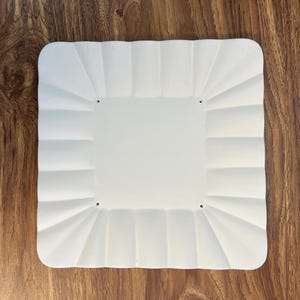 May include: A square white plate with rounded corners and a scalloped, fluted border design. The center of the plate is flat and smooth, featuring four small, evenly spaced holes positioned near each corner of the central square section.