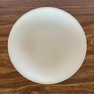 May include: A circular, off-white ceramic bowl with a smooth, matte finish. The interior surface of the bowl is plain and features a single, small dark speck near the center, showcasing a simple, functional design for serving food or display.