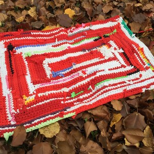 May include: A red, white, and multi-colored crocheted rug with a square design. The rug is lying on a bed of brown leaves.