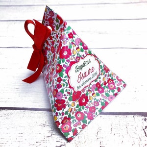 Puede incluir: Una caja blanca en forma de pirámide con una cinta roja y un patrón floral. La caja tiene una etiqueta que dice "Baptême Isaura 1er décembre 2018".