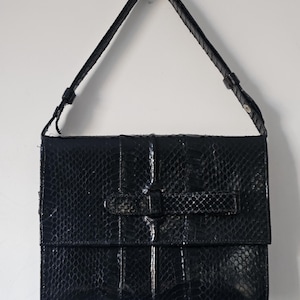 May include: A black handbag with a glossy, textured snakeskin pattern. The bag features a rectangular flap closure secured by a horizontal strap detail across the centre. It includes a single adjustable shoulder strap attached to the top sides.