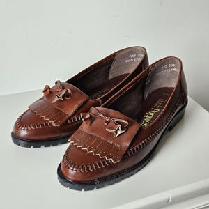 May include: A pair of brown leather loafers featuring a fringed kiltie tongue, a decorative bow, and a moccasin-style stitched toe. The interior lining is printed with the text Hush Puppies and size information 110 200 Size 5 2727.