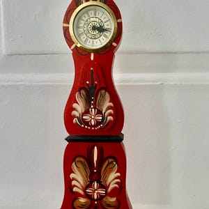 May include: A red wooden clock with a gold-rimmed clock face. The clock features floral designs in brown, tan, and white. The clock is approximately 30 cm tall and has a black base.