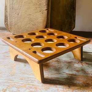 May include: A wooden wine glass holder with a square top and rounded holes for glasses. The holder is made of light brown wood and has a sturdy base. Two wooden cutting boards are in the background.