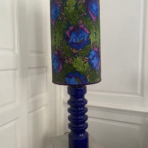 Puede incluir: Una lámpara de pie de cerámica azul con una pantalla de lámpara con estampado floral. La pantalla tiene un ribete de color morado oscuro y presenta flores azules, moradas y verdes sobre un fondo verde oscuro.