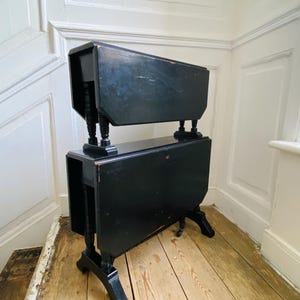May include: Two black antique drop-leaf tables stacked. The tables have turned legs and a distressed finish. The tables are positioned in a corner with light-coloured wooden floors and white walls. The tables are approximately 76 cm high.