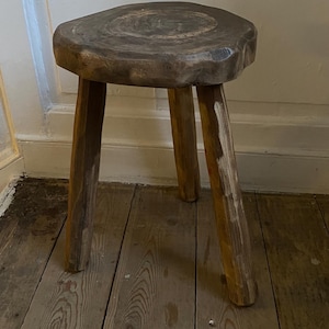 May include: A rustic, three-legged wooden stool with a round, textured seat. The stool is made of unfinished wood, showcasing natural grain patterns and knots. The legs are sturdy and angled, providing stability. The stool is a natural brown colour.