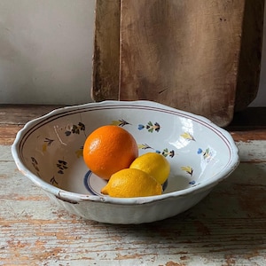 May include: A white, oval ceramic bowl with a scalloped edge, adorned with blue and yellow floral designs, contains an orange and two lemons. The bowl rests on a distressed wooden surface, with two wooden chopping boards behind.