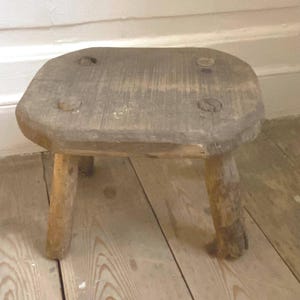 May include: A small, rustic wooden stool. It has a rectangular seat and three sturdy legs. The stool is made of weathered wood, showing natural grain and knots. The legs are slightly tapered, and the design suggests a handcrafted piece.
