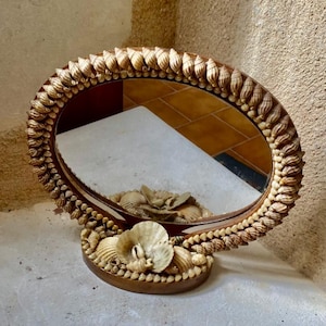 Puede incluir: Espejo ovalado con marco y base adornados con conchas marinas. El marco está bordeado con pequeñas conchas puntiagudas, y la base presenta conchas más grandes. El espejo refleja el entorno.