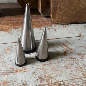 May include: Three stainless steel piping tips of varying sizes, each with a black base, are arranged on a distressed, light-colored wooden surface. The tips are cone-shaped, with a small opening at the top. The background includes wooden cutting boards.