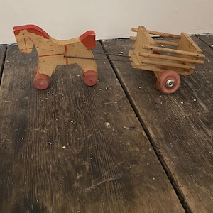 May include: A vintage wooden toy horse pulling a small wagon. The horse is tan with red accents on its mane, tail, and wheels. The wagon is made of light-colored wood with red wheels. The toy is connected by a metal wire.