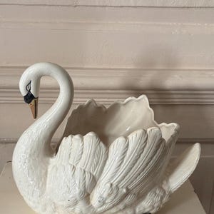 May include: A white ceramic swan-shaped planter or bowl featuring a glossy, crackled glaze finish. The swan has a long, curved neck, a black and gold beak, and detailed moulded feather textures across its body, with a hollow centre for holding items.