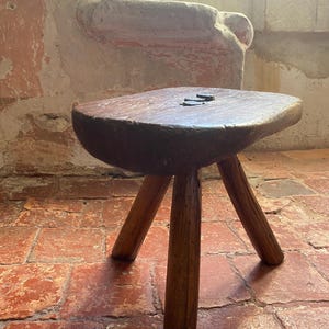 May include: A small, three-legged wooden stool with a thick, dark brown, irregularly shaped seat and three sturdy, cylindrical legs. The seat features a visible grain and two small, dark metal fasteners embedded in the top surface.