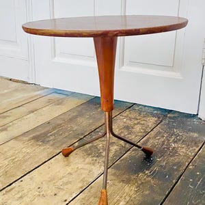 Puede incluir: Una pequeña mesa auxiliar de estilo moderno de mediados de siglo con una tapa redonda de madera. La mesa tiene un pedestal ensanchado de madera marrón y tres patas de metal. La mesa mide aproximadamente 61 cm de altura.
