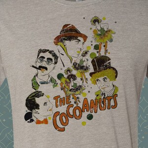 May include: A heather grey t-shirt with a vintage-style illustration of a group of people with the text "The Cocoanuts" printed on the front.