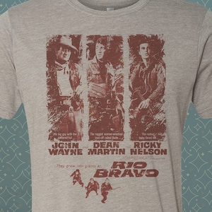 May include: A heather grey t-shirt with a vintage-style print of John Wayne, Dean Martin, and Ricky Nelson from the movie "Rio Bravo". The text reads "The big guy with the powerful hat, John Wayne. The rugged somewhat wrecked part of faded Duke, Dean Martin. The reckless, fully blood red, Ricky Nelson. They grew into giants at Rio Bravo."