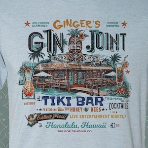 May include: Light blue t-shirt with a graphic design of a tiki bar called "Ginger's Gin Joint" featuring a tropical scene with palm trees, cocktails, and the text "Tiki Bar Tropical Cocktails Featuring The Honey Bees Live Entertainment Nightly Honolulu, Hawaii 34 Tiki Pop Trading Co."