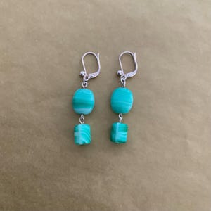 Può includere: Orecchini pendenti con ganci color argento. Ogni orecchino presenta due perline turchesi: una ovale e una quadrata. Le perline hanno un motivo a righe e sono collegate da piccoli anelli color argento. Un accessorio elegante.