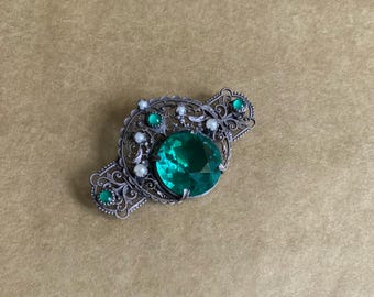 May include: A silver filigree brooch with a large green gemstone surrounded by smaller green and white gemstones.