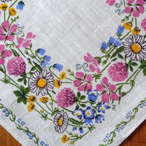 Könnte beinhalten: Weißes Leinen mit floralem Muster. Das Design zeigt rosa Klee, Gänseblümchen, blaue Vergissmeinnicht und gelbe Blumen. Ein Rand aus blauen Blumen umrahmt den Stoffrand.