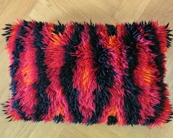 May include: A rectangular throw pillow featuring a thick, shaggy pile texture. The surface displays a repeating vertical striped pattern in alternating bands of deep black, vibrant magenta, and bright orange yarn fibers throughout the entire piece.