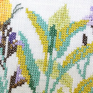 Puede incluir: Primer plano de un dise&ntilde;o floral de punto de cruz con flores amarillas, verde claro y moradas con tallos y hojas marrones. La labor de aguja est&aacute; sobre un fondo de tela blanca.